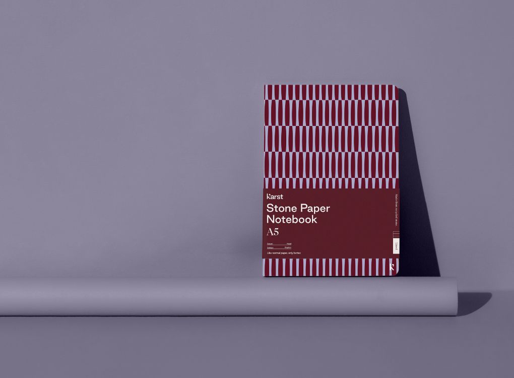 A Studio Muse A5 hardcover notebook by Karst with a maroon and lavender geometric patterned cover, standing against a lavender background. The notebook features smooth, recycled stone paper with lined pages and a slim, lightweight cover.