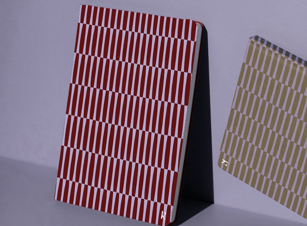 Two Studio Muse hardcover notebooks leaning against a lavender wall. The notebook on the left has a maroon and lavender geometric pattern, while the one on the right features an olive green and lavender pattern. Both have slim, lightweight covers with a repeating vertical stripe design.