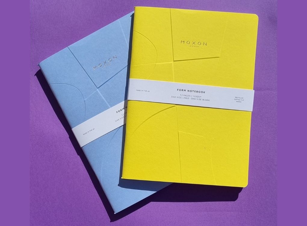 Yellow and blue MOXON B5 ‘Form’ notebooks stacked with packaging bands on a purple background