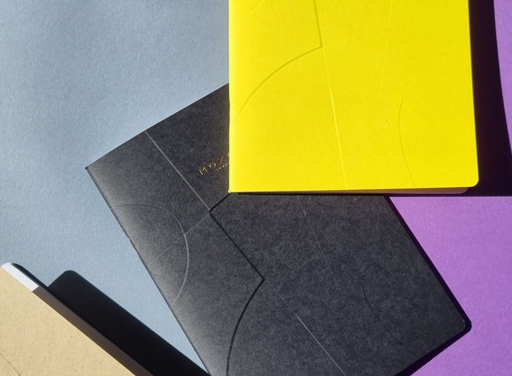 Black MOXON B5 ‘Form’ notebook placed over kraft, blue, and yellow notebooks on purple background