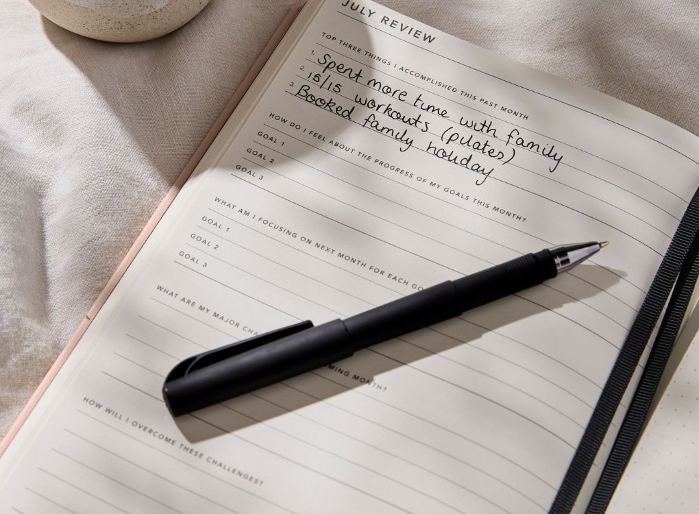 MiGoals 2026 Classic Diary open to a July review spread with handwritten notes about family, workouts, and holidays, styled with a black pen and natural fabric backdrop.