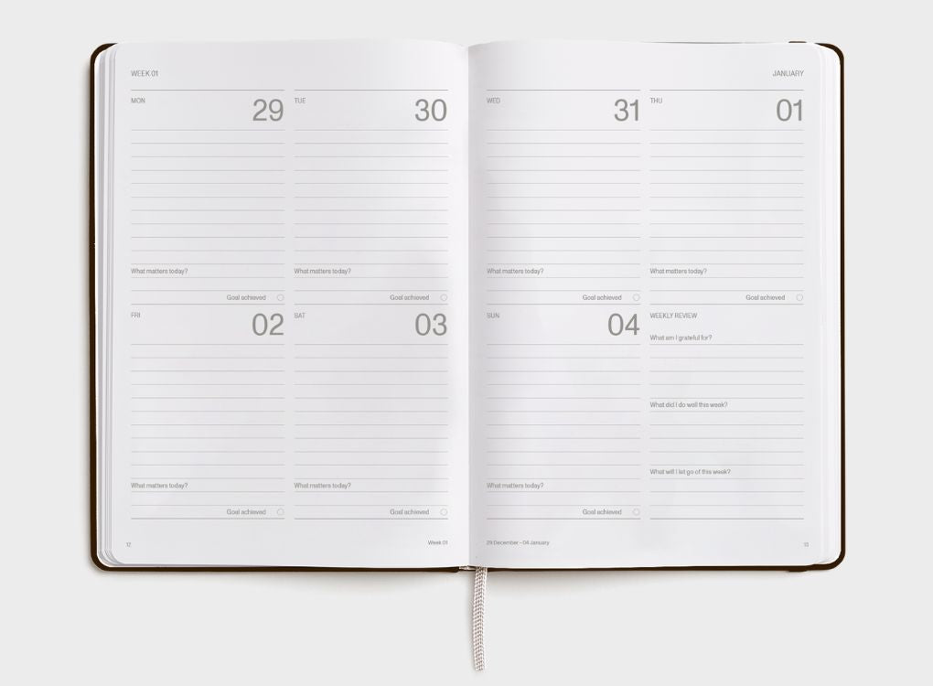 A hand holding a brown 2026 Karst weekly planner against a dark background. The planner has minimalist white text displaying "2026" in the center and "WEEKLY PLANNER" at the bottom.