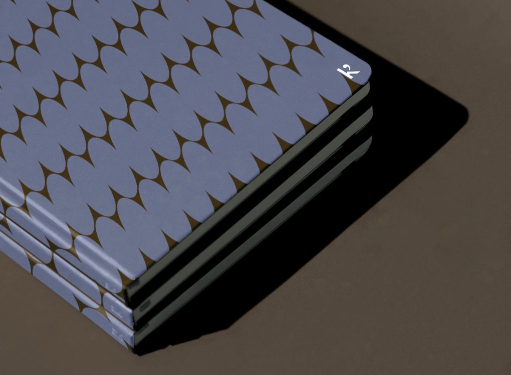 Three stacked Studio Muse hardcover notebooks by Karst, each with a blue and brown abstract checkerboard patterned cover, lying flat on a dark brown surface with a strong shadow cast to the right. The notebooks feature smooth, recycled stone paper and slim covers.