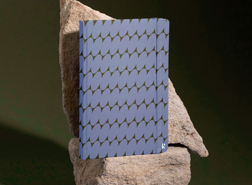 A Studio Muse hardcover notebook by Karst with a blue and brown abstract checkerboard patterned cover, standing upright against a textured beige stone and a dark green background. The notebook features smooth, recycled stone paper and a slim cover with an elastic band.