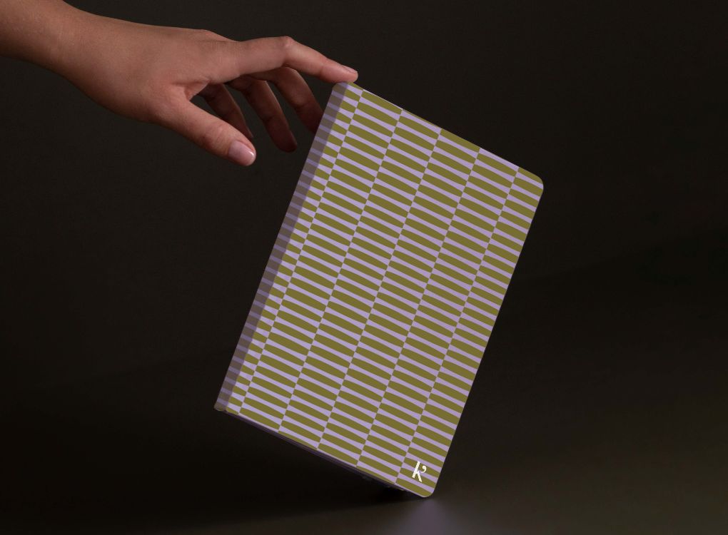 A hand with natural nails reaching toward a Studio Muse hardcover notebook by Karst, featuring an olive green and lavender geometric patterned cover. The notebook is held upright against a dark background and has a slim, lightweight cover with recycled stone paper.
