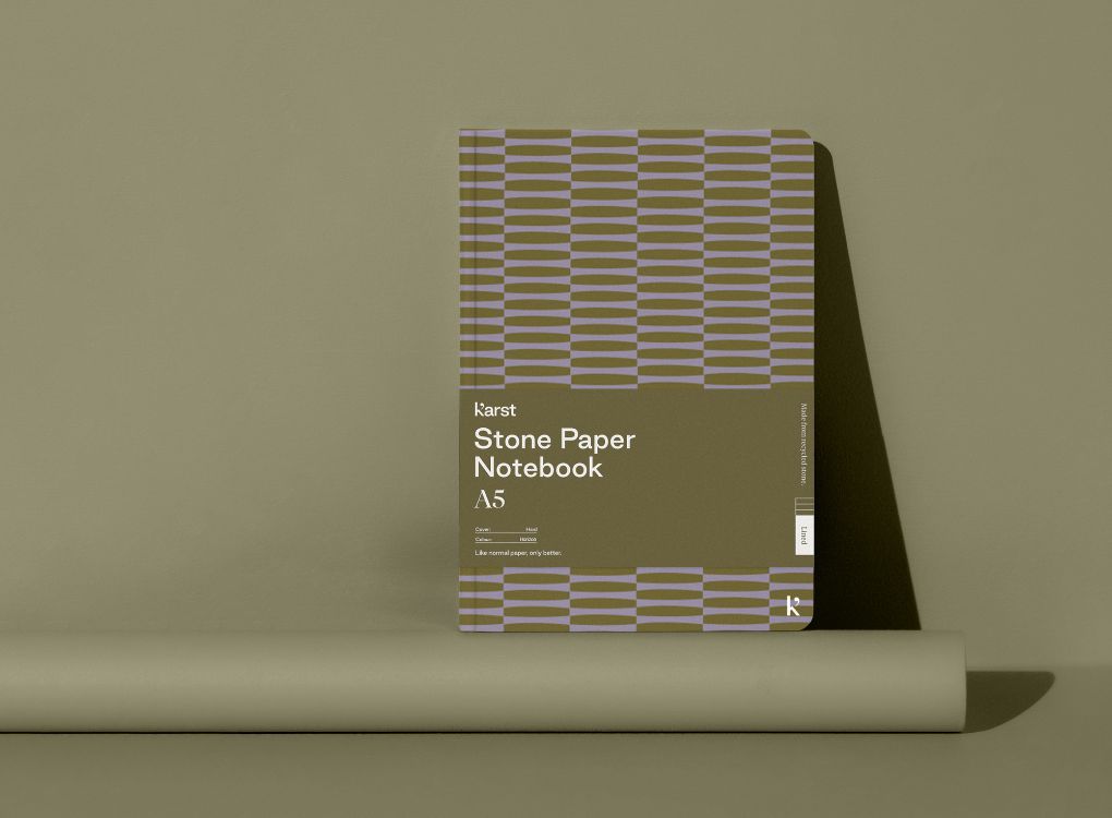A Studio Muse A5 hardcover notebook by Karst with an olive green and lavender geometric patterned cover, standing against a matching olive green background. The notebook features smooth, recycled stone paper with lined pages and a slim, lightweight cover.