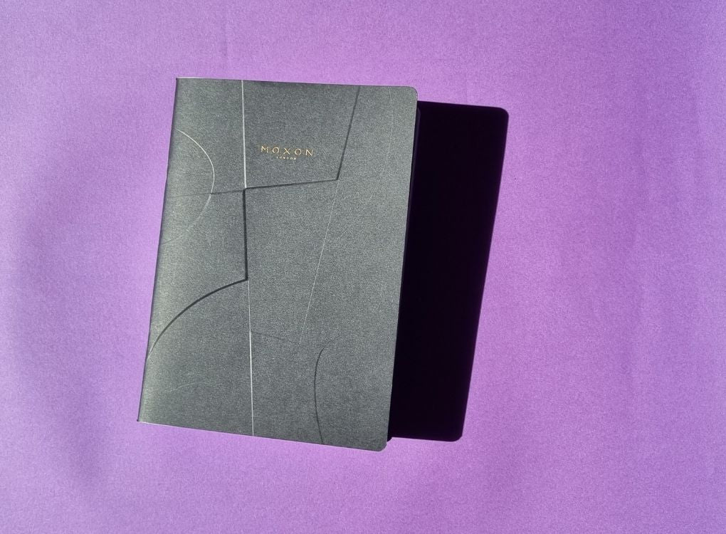 Black MOXON B5 ‘Form’ notebook on a purple surface with sunlight and shadow