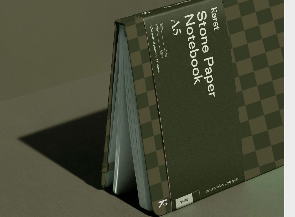 A Studio Muse A5 hardcover notebook by Karst with a dark olive green checkerboard patterned cover, standing open on a matching olive green surface. The notebook features smooth, recycled stone paper with lined pages and a slim, lightweight cover with an elastic band.