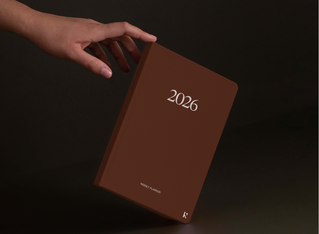 A hand holding a brown 2026 Karst weekly planner against a dark background. The planner has minimalist white text displaying "2026" in the center and "WEEKLY PLANNER" at the bottom.