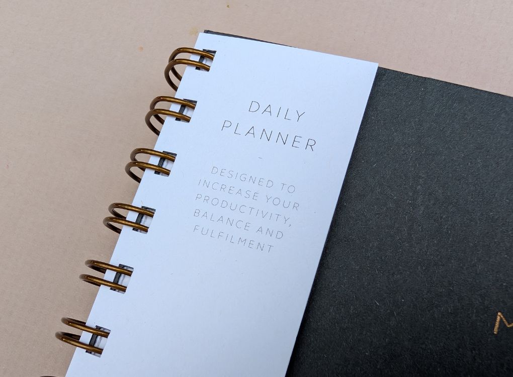 MOXON Daily planner with gold spiral binding on a beige background. Shows info tab on the front of the planner