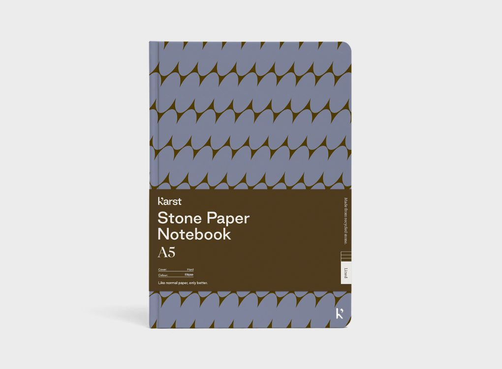 A Studio Muse A5 hardcover notebook by Karst with an olive green and lavender geometric patterned cover, standing against a matching olive green background. The notebook features smooth, recycled stone paper with lined pages and a slim, lightweight cover.